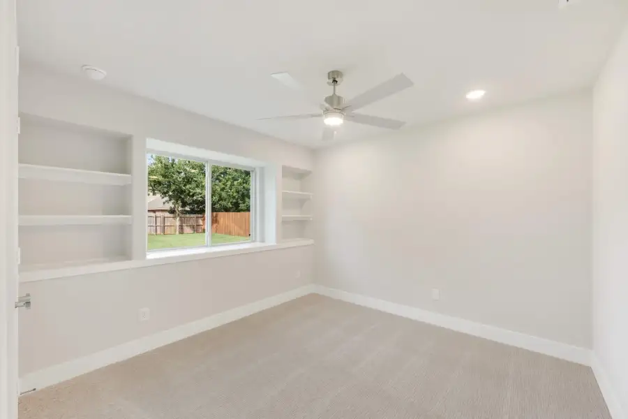 Empty bedroom with built-in shelves, ceiling fan, carpet flooring, and a window overlooking a backyard