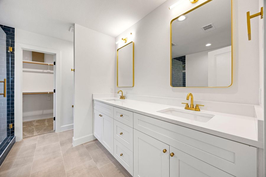 Luxurious primary bathroom featuring a double-sink vanity with quartz countertops, white cabinetry, and elegant gold fixtures. Large framed mirrors, recessed lighting, and a clean modern design create a spa-like atmosphere with direct access to the walk-in closet.