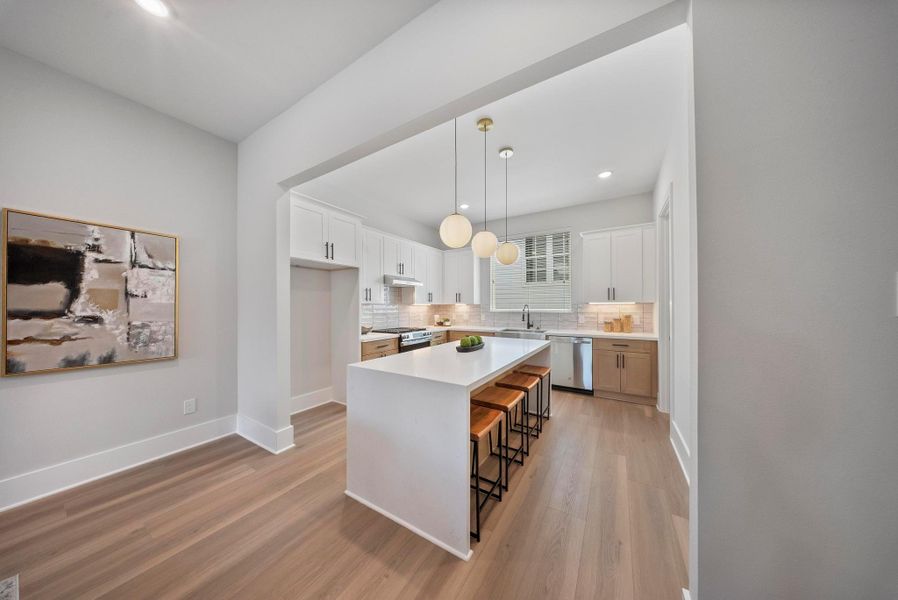 This photo showcases a modern kitchen with a spacious, open layout. It features a large island with seating, sleek white cabinetry, stainless steel appliances, and stylish pendant lighting. The light wood flooring and neutral color palette create a warm and inviting atmosphere.