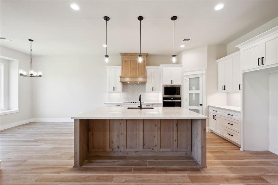 Kitchen with hanging light fixtures, custom range hood, white cabinetry, and a kitchen island with sink