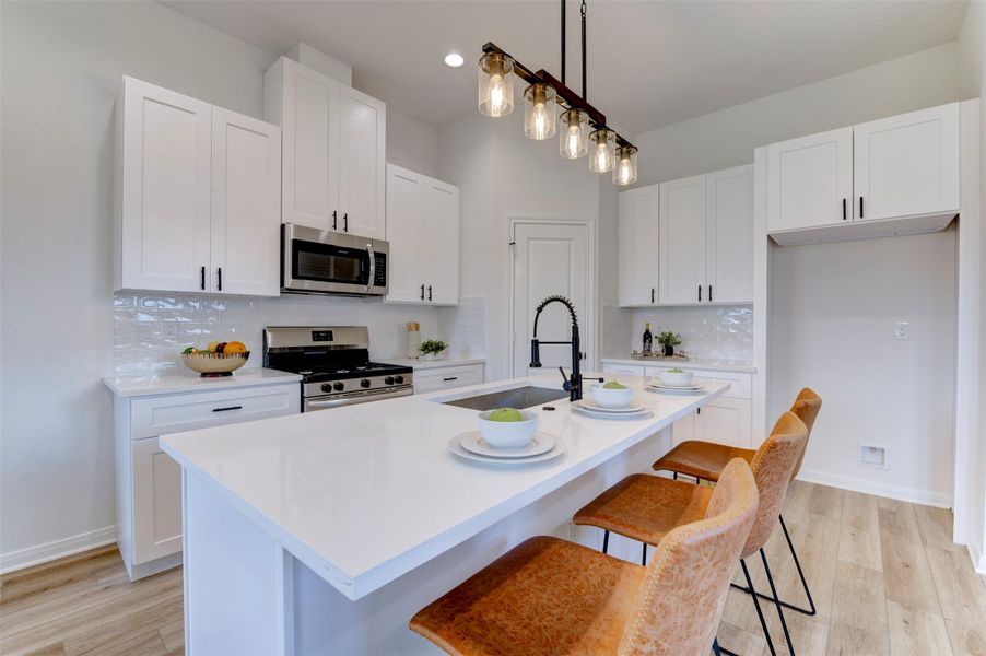 Kitchen with white shaker cabinets, quartz countertops, subway tile backsplash, stainless gas range, stainless microwave, farmhouse sink on island, matte black coil-spring faucet, Edison rail pendant lighting, and leather bar stools. Pantry door visible at rear.