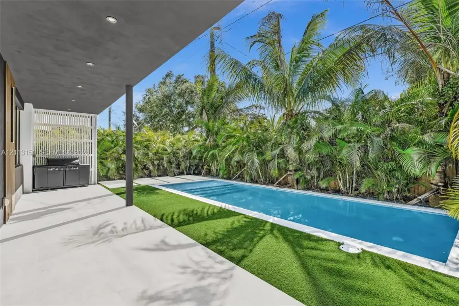 Covered patio with recessed lighting, rectangular swimming pool, integrated built-in outdoor kitchen, synthetic turf, and privacy-screening foliage Covered patio with recessed lighting, rectangular swimming pool, integrated built-in outdoor kitchen, synthetic turf, and privacy-screening foliage