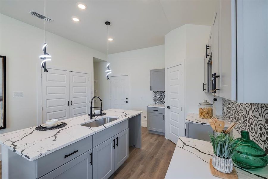 Kitchen with sink, tasteful backsplash, light stone counters, decorative light fixtures, and a center island with sink