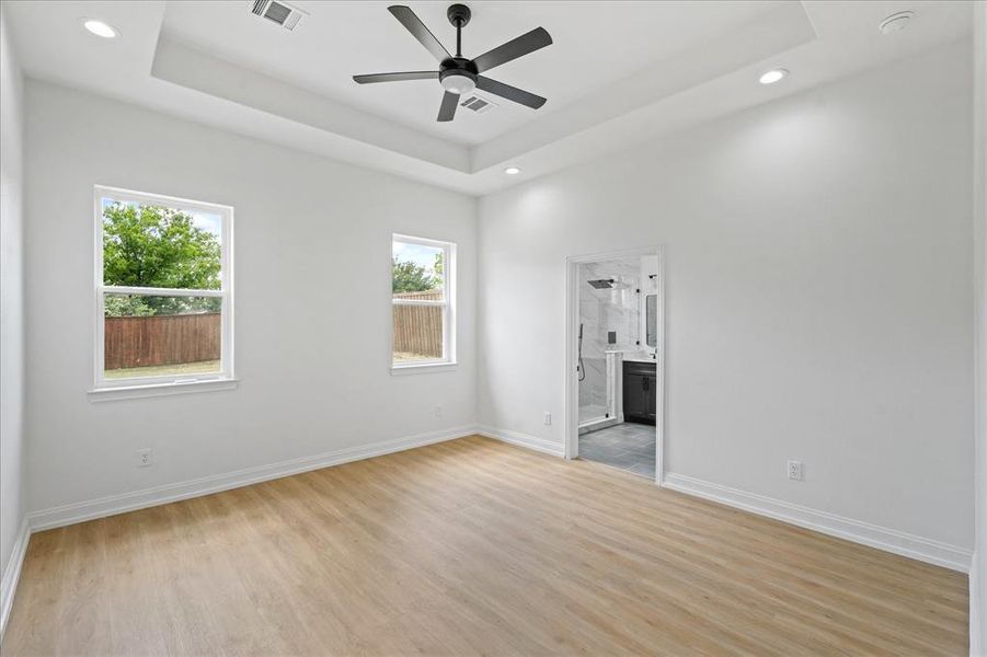 Spacious Primary Bedroom featuring light wood-finish flooring, a tray ceiling with a ceiling fan, recessed lighting, and dual windows