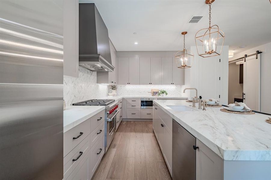 Kitchen featuring backsplash, appliances with stainless steel finishes, an island with sink, light wood-style flooring, and a barn door Kitchen featuring backsplash, appliances with stainless steel finishes, an island with sink, light wood-style flooring, and a barn door