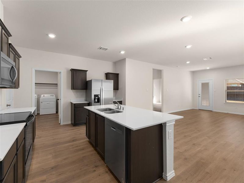 Kitchen with washing machine and clothes dryer, sink, wood-type flooring, appliances with stainless steel finishes, and a kitchen island with sink