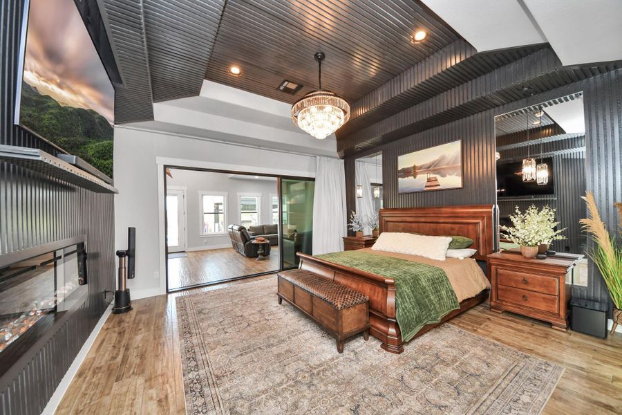 Luxurious bedroom featuring a stylish tray ceiling, elegant chandelier, and a cozy fireplace. The space opens into a bright sitting area, enhancing the room's spaciousness and inviting ambiance.