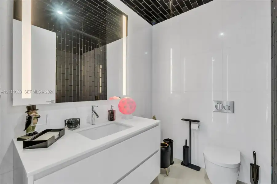 Modern floating vanity featuring a rectangular integrated sink and white stone-finish countertop, complemented by a large rectangular mirror with integrated LED lighting, wall-mounted dual-flush toilet system, and high-contrast black subway tile detailing on the ceiling and wall Modern floating vanity featuring a rectangular integrated sink and white stone-finish countertop, complemented by a large rectangular mirror with integrated LED lighting, wall-mounted dual-flush toilet system, and high-contrast black subway tile detailing on the ceiling and wall