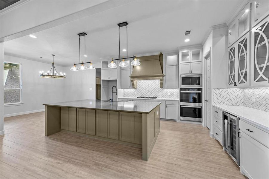 Kitchen with custom exhaust hood, a kitchen island with sink, hanging light fixtures, wine cooler, and light wood-type flooring