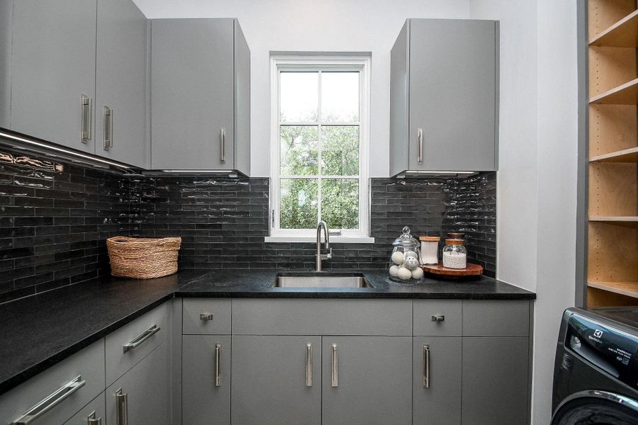 This is a modern laundry room with sleek gray cabinets, black countertops, and a black tiled backsplash. It features a stainless steel sink under a window that provides natural light. The space is organized, with open shelving and room for appliances.