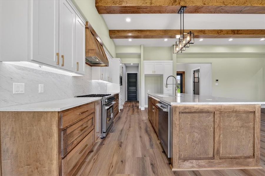 Modern kitchen featuring dual-tone cabinetry, a central island with a built-in sink, stainless steel appliances, wood-finish flooring, and exposed ceiling beams