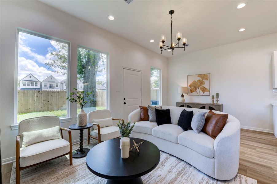 Open-concept view from living room through to kitchen and dining, showing hardwood floors, high ceilings, recessed lighting, dual pendant chandeliers, white kitchen with island bar seating, and stainless appliances.