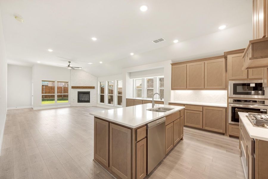 Open kitchen with large island, tan wood cabinets, stainless appliances, and light hardwood floors open to living room with fireplace. Open kitchen with large island, tan wood cabinets, stainless appliances, and light hardwood floors open to living room with fireplace.