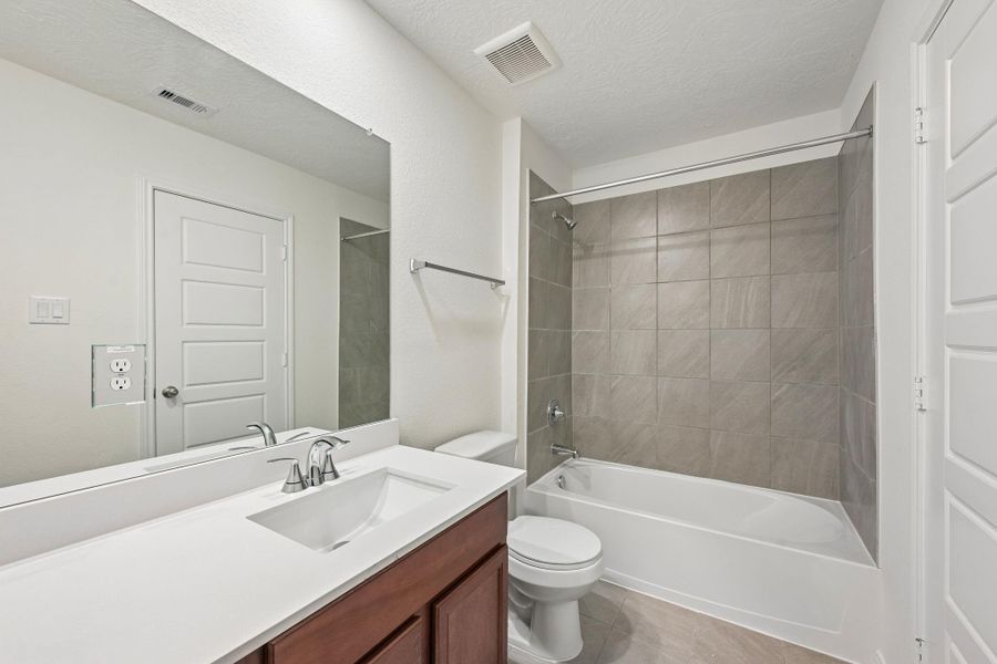 The secondary bathroom offers tile flooring, a tub/shower combination with a stylish tile surround, rich stained wood cabinetry with added knee space and elegant light countertops, a mirror, and sleek fixtures with modern finishes.
