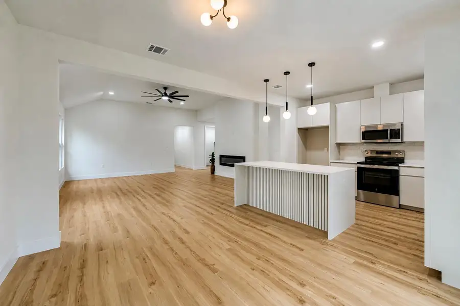 Open-concept living space featuring wood-finish flooring, a modern kitchen with a white island and pendant lighting, stainless steel appliances, and a built-in electric fireplace