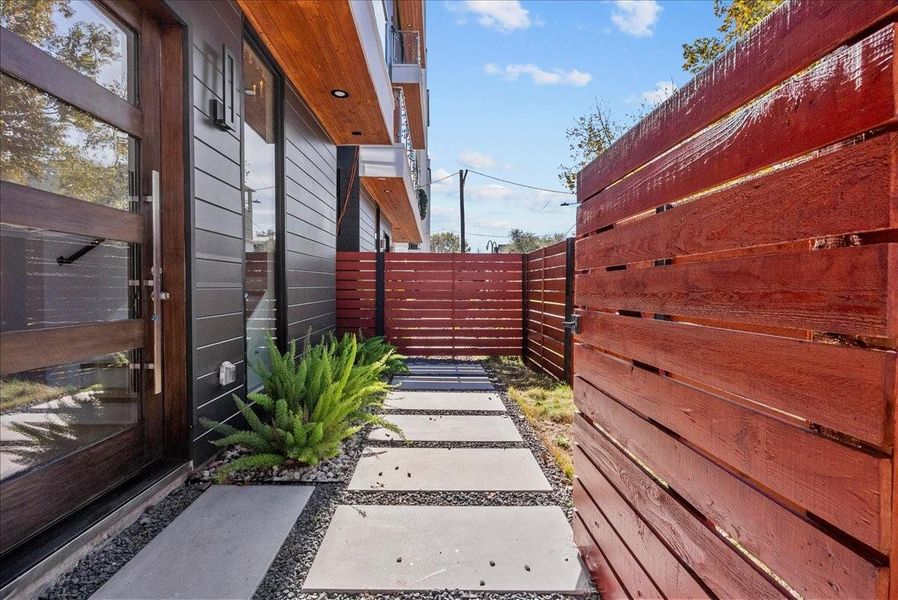 This photo showcases a modern side yard with a sleek wooden fence and a pathway of concrete slabs surrounded by gravel. The exterior features dark siding with a glass door and a small garden area with lush greenery, offering a clean and contemporary outdoor space. This photo showcases a modern side yard with a sleek wooden fence and a pathway of concrete slabs surrounded by gravel. The exterior features dark siding with a glass door and a small garden area with lush greenery, offering a clean and contemporary outdoor space.