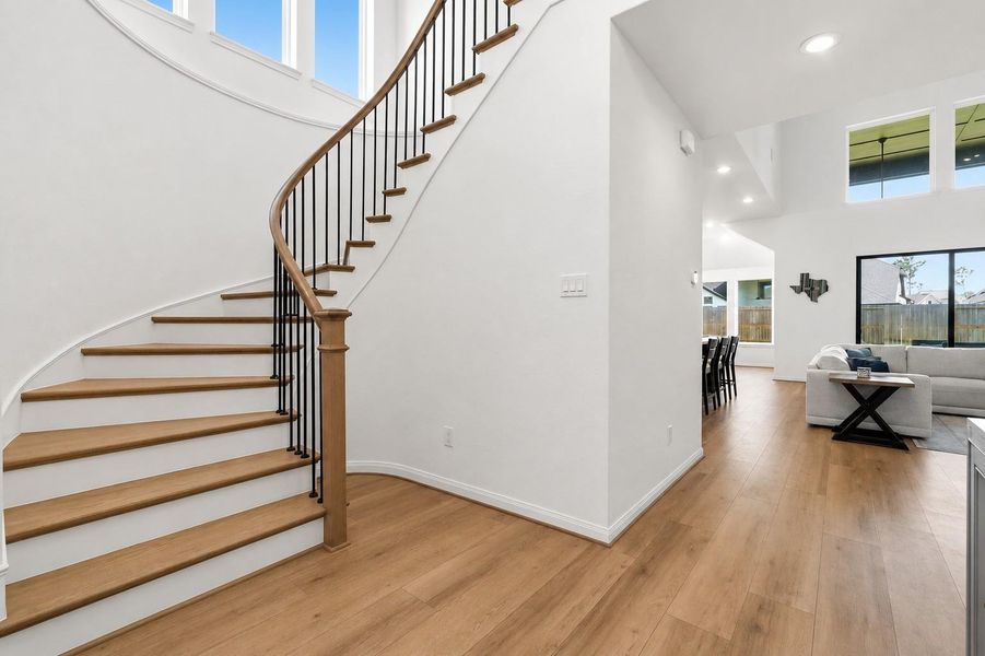 This photo showcases a modern, open-concept living area with a curved staircase featuring wooden steps and black railing. The space is bright with natural light from large windows, and the living room and dining area flow seamlessly with wood-style vinyl plank flooring throughout.