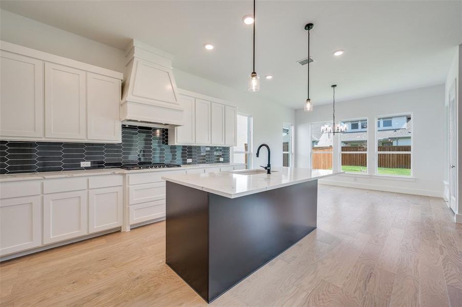 Kitchen featuring decorative light fixtures, decorative backsplash, custom range hood, light hardwood / wood-style floors, and a kitchen island with sink