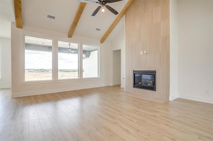 Unfurnished living room featuring a tiled fireplace, light wood-type flooring, high vaulted ceiling, beamed ceiling, and ceiling fan