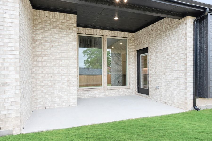 Covered back patio with light brick walls, dark ceiling, concrete floor, and black-framed glass door leading inside