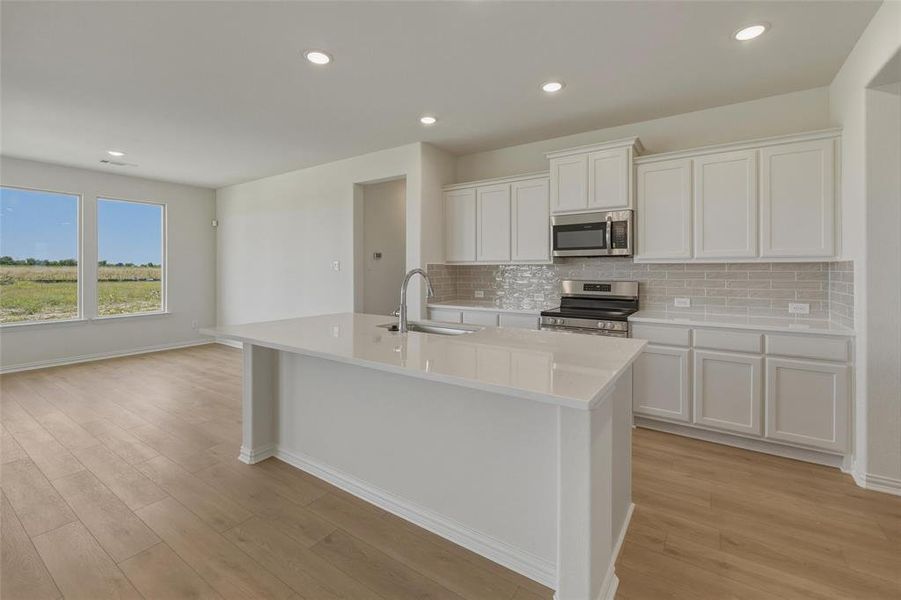Kitchen island with a built-in sink and stainless steel faucet, white cabinetry, integrated stainless steel microwave, gas range, light-toned subway tile backsplash, and recessed lighting Kitchen island with a built-in sink and stainless steel faucet, white cabinetry, integrated stainless steel microwave, gas range, light-toned subway tile backsplash, and recessed lighting