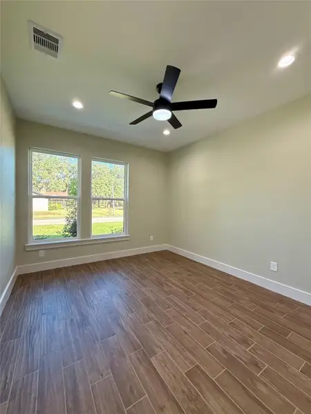 Bright room with wood-like tile flooring, beige walls, and two large windows offering natural light. Features a modern ceiling fan and recessed lighting, creating a cozy and inviting atmosphere.