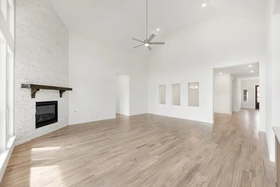 Open living room with vaulted ceiling, white stone fireplace, wood mantel, and light hardwood floors Open living room with vaulted ceiling, white stone fireplace, wood mantel, and light hardwood floors