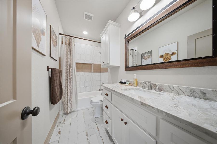 Bathroom featuring a white vanity with a stone countertop and an integrated sink, a rectangular framed mirror with overhead lighting, a white bathtub with a tiled surround, wood-finish flooring, and a built-in wall cabinet