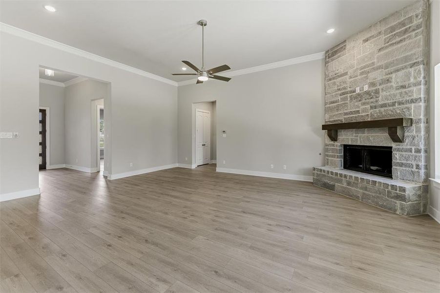 Spacious living area featuring a stone-clad fireplace with a dark wood mantel, light wood-finish flooring, a ceiling fan with integrated lighting, recessed lighting, and crown molding