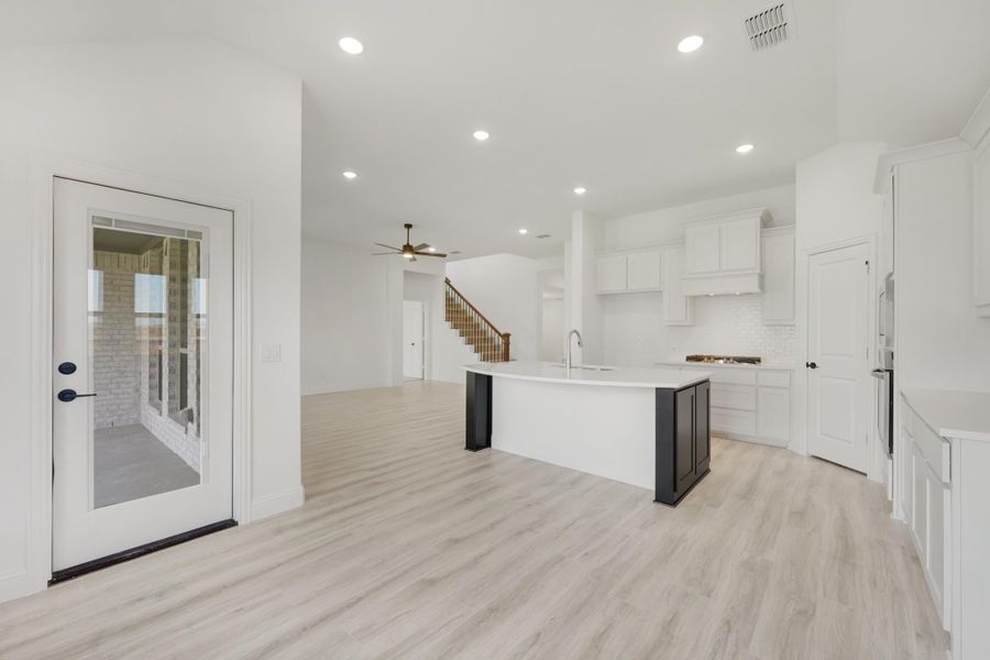 Open-concept kitchen and living area with white cabinets, curved island, light wood floors, and staircase in background.