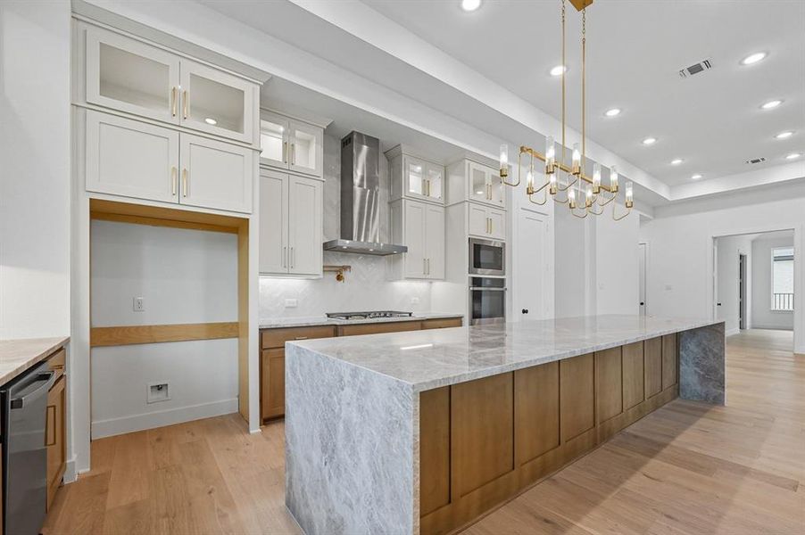 Kitchen featuring light stone counters, glass insert cabinets, a large island, light wood-style floors, and wall chimney exhaust hood Kitchen featuring light stone counters, glass insert cabinets, a large island, light wood-style floors, and wall chimney exhaust hood