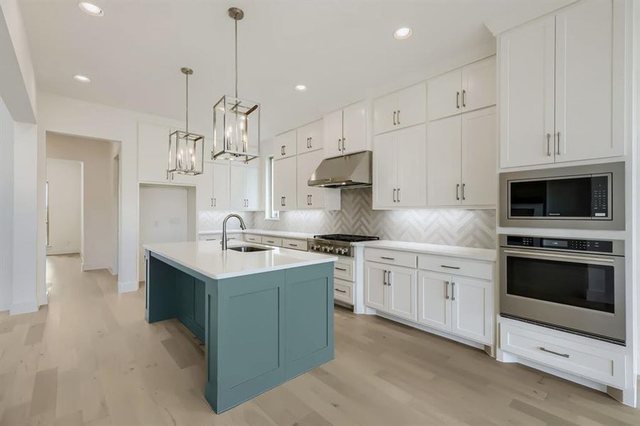 Modern kitchen featuring light wood-finish flooring, white upper and lower cabinetry, a blue-painted kitchen island with a white countertop, stainless steel appliances, and two hanging pendant light fixtures Modern kitchen featuring light wood-finish flooring, white upper and lower cabinetry, a blue-painted kitchen island with a white countertop, stainless steel appliances, and two hanging pendant light fixtures