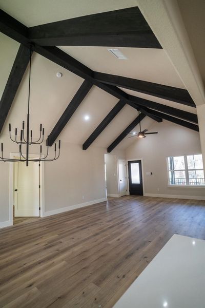 Unfurnished living room featuring beam ceiling, high vaulted ceiling, ceiling fan with notable chandelier, wood finished floors, and baseboards