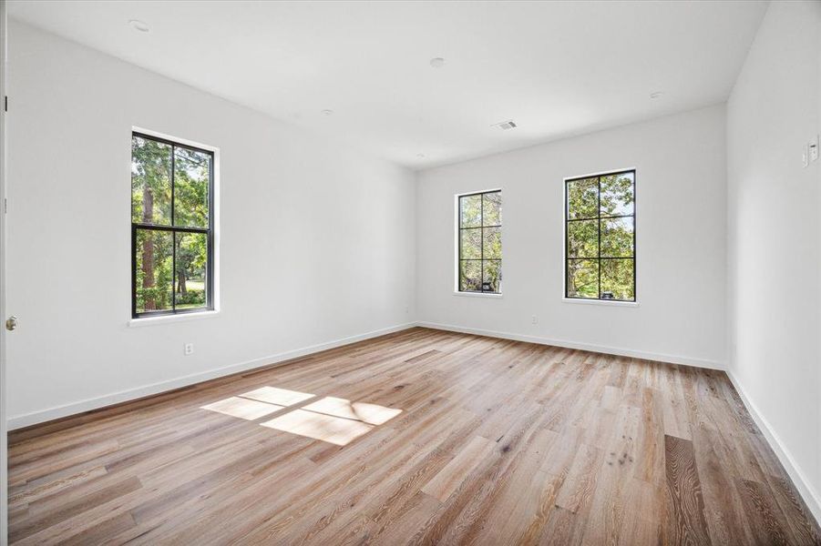 This spacious bedroom is filled with natural light from three large windows, offering a tranquil view of the surrounding greenery. The European oak wood flooring adds warmth to the room, while the simple, white walls provide a clean, modern backdrop. The layout is versatile, ready to accommodate various furniture arrangements.