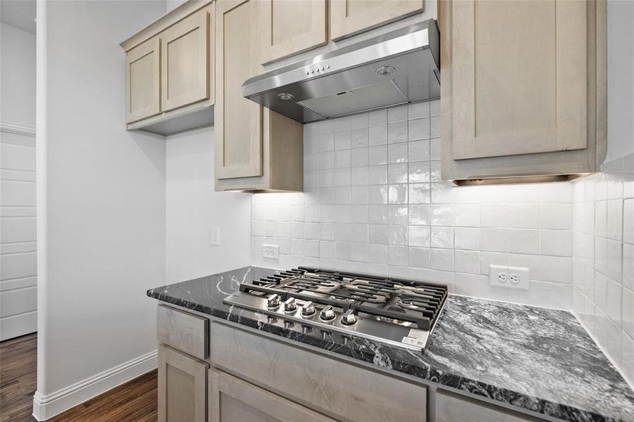 Kitchen featuring dark stone countertops, tasteful backsplash, under cabinet range hood, and light brown cabinets