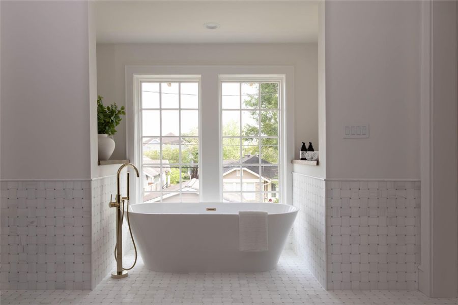 Spa-inspired primary bath with a freestanding soaking tub centered beneath a double Pella window with neighborhood views. Floor-to-ceiling basketweave marble tile on walls and floors, brass floor-mount faucet with handheld attachment, and built-in marble ledges on both sides of the tub alcove. Spa-inspired primary bath with a freestanding soaking tub centered beneath a double Pella window with neighborhood views. Floor-to-ceiling basketweave marble tile on walls and floors, brass floor-mount faucet with handheld attachment, and built-in marble ledges on both sides of the tub alcove.