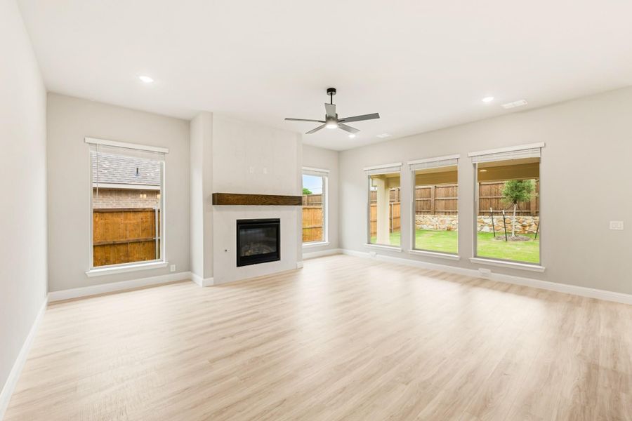 Open living room with light wood floors, fireplace with wood mantel, ceiling fan, and large windows overlooking backyard Open living room with light wood floors, fireplace with wood mantel, ceiling fan, and large windows overlooking backyard