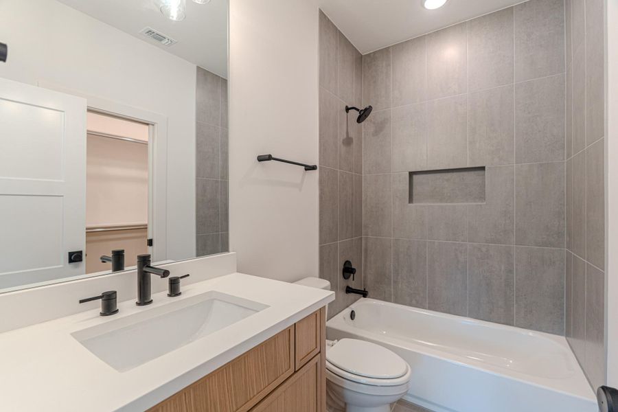 This modern bathroom features a sleek vanity with a white countertop and black fixtures. The bathtub has a gray tiled surround with a built-in niche for storage, and there's a closet visible through a sliding door.