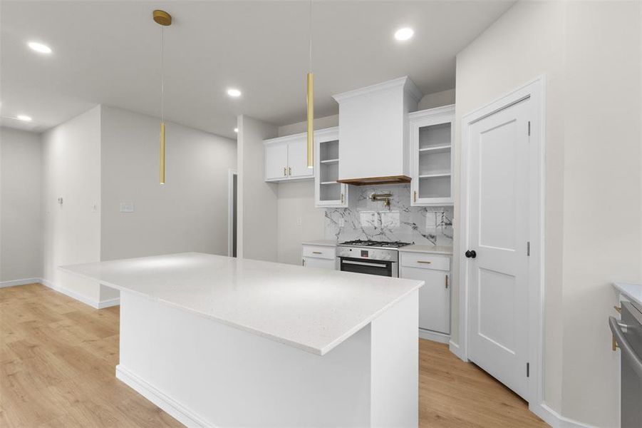 Spacious kitchen island featuring a light-toned countertop with integrated sink, gold-toned pendant lighting, wood-finish flooring, white cabinetry with glass-front uppers, and a marble-look backsplash