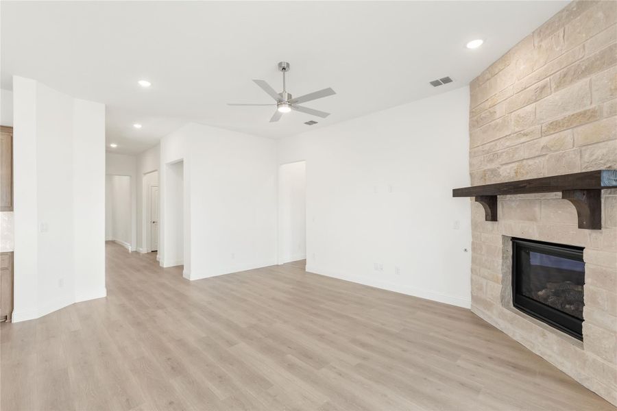 Open living room with light wood floors, ceiling fan, and stone fireplace with dark wood mantel Open living room with light wood floors, ceiling fan, and stone fireplace with dark wood mantel