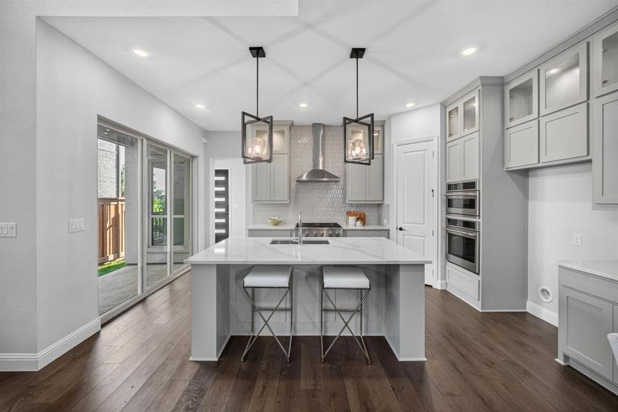 Modern kitchen featuring a central island with a sink and white countertop, light gray cabinetry, stainless steel appliances, and wood-finish flooring