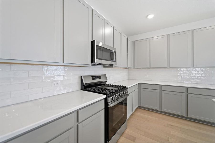 Contemporary kitchen featuring light gray cabinetry, white countertops, a subway tile backsplash, stainless steel appliances, and light wood-finish flooring Contemporary kitchen featuring light gray cabinetry, white countertops, a subway tile backsplash, stainless steel appliances, and light wood-finish flooring