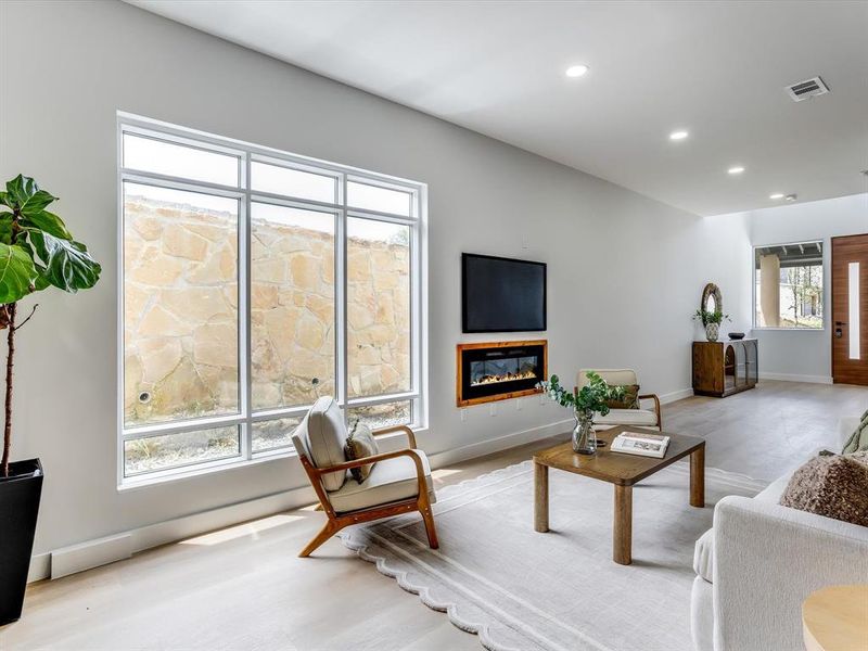 Spacious interior featuring large picture windows, wood-finish flooring, a wall-mounted electric fireplace, and recessed ceiling lighting Spacious interior featuring large picture windows, wood-finish flooring, a wall-mounted electric fireplace, and recessed ceiling lighting