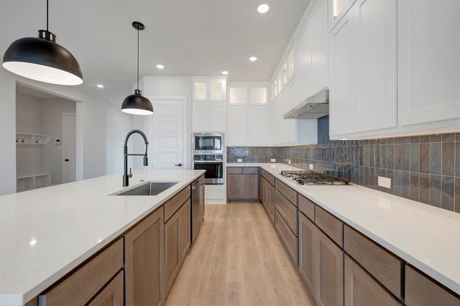 Kitchen with light wood-style floors, light stone countertops, two tone cabinets, hanging light fixtures, and stainless steel appliances