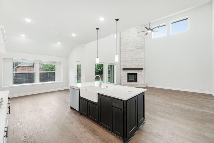 Open-concept kitchen with dark island, farmhouse sink, and tall stone fireplace in living area with vaulted ceiling Open-concept kitchen with dark island, farmhouse sink, and tall stone fireplace in living area with vaulted ceiling