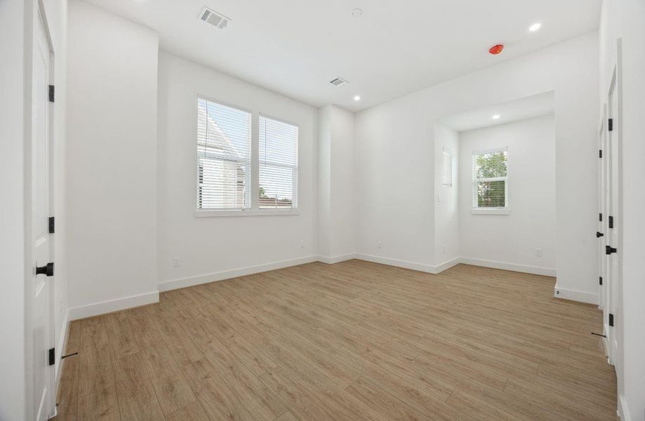 Spacious primary bedroom with high ceilings, recessed lighting, warm wood-look flooring, and multiple windows that fill the room with natural light. The layout includes a defined nook that adds flexibility for a sitting area, desk, reading corner, or additional furniture while maintaining a clean, open feel.