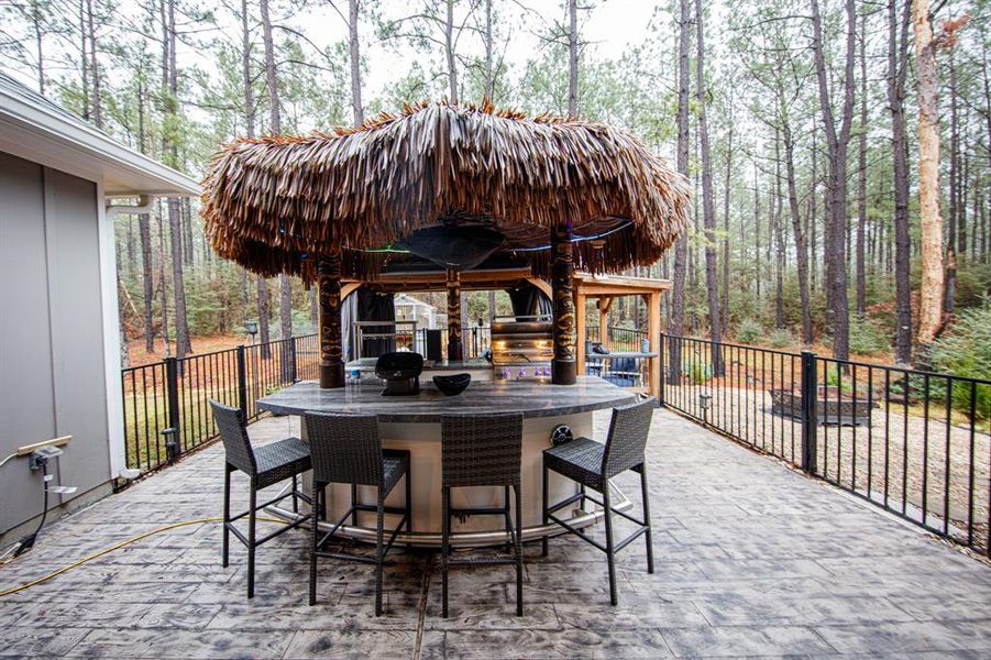 This photo showcases a charming outdoor patio area featuring a tiki-style bar, grill, wet bar, wine fridge etc. topped with a thatched roof. Surrounded by trees, the space offers a serene, wooded backdrop, perfect for entertaining or relaxing. The bar includes seating and is enclosed by a decorative railing, adding a cozy and inviting atmosphere to the property.