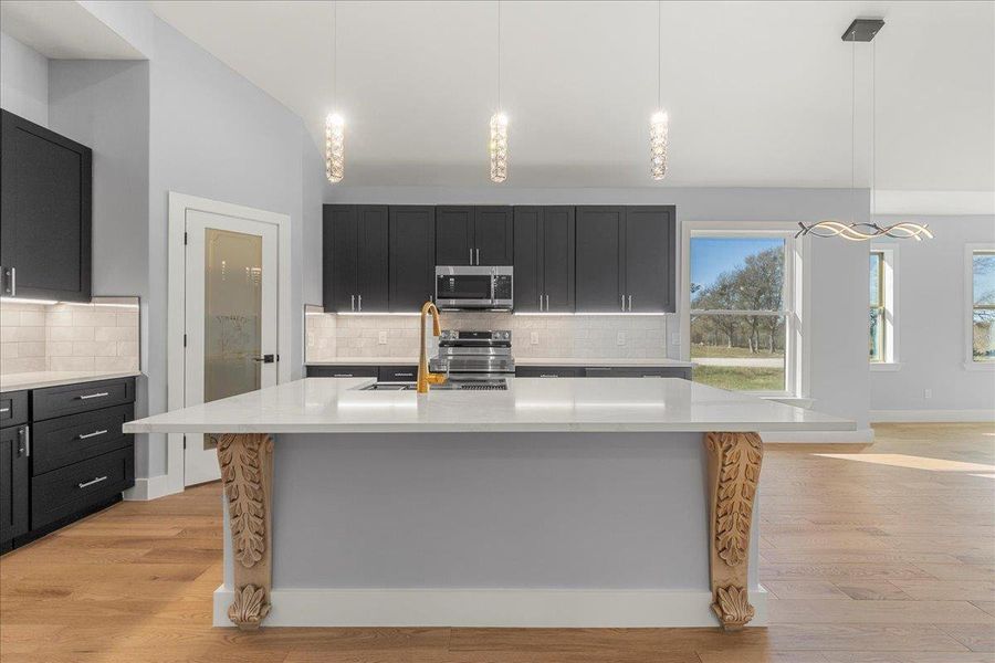 Kitchen with hanging light fixtures, light stone counters, a center island with sink, stainless steel appliances, and dark cabinets Kitchen with hanging light fixtures, light stone counters, a center island with sink, stainless steel appliances, and dark cabinets