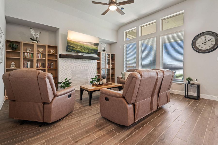 Bright and inviting living room with large windows, modern ceiling fan, and cozy recliner seating. Features a stylish fireplace with built-in shelves and a wall-mounted TV. Perfect for relaxing and entertaining. Bright and inviting living room with large windows, modern ceiling fan, and cozy recliner seating. Features a stylish fireplace with built-in shelves and a wall-mounted TV. Perfect for relaxing and entertaining.