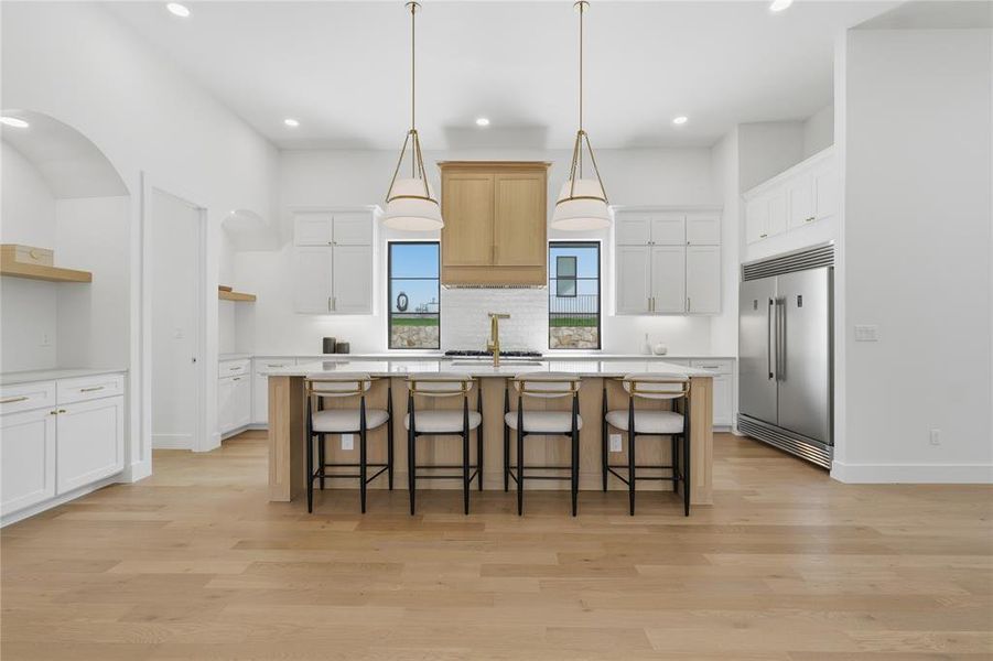 Gourmet kitchen featuring a central island with bar seating, stainless steel appliances, extensive white cabinetry, light wood-finish flooring, and two statement pendant lights